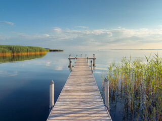 Obraz premium Wooden pier extends into a calm lake, surrounded by reeds and open water under a partly cloudy sky. The tranquil waterfront scene features strong leading lines, reflections, and ample copy space.