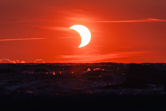 View of a partial solar eclipse over the dark sea at sunset with a vibrant red sky in Merikula, Harju maakond, Estonia.