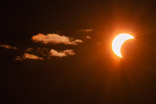 View of a partial solar eclipse with the sun partially obscured by the moon, glowing orange against a dark sky with wispy clouds in Merikula, Harju maakond, Estonia.