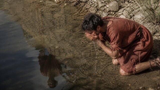 Repentant young man kneeling beside a river and washing his face. Biblical prodigal son seeking renewal, humility, forgiveness, and spiritual transformation.