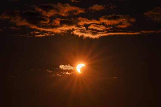 View of a partial solar eclipse with the sun partially obscured by the moon, surrounded by dark clouds and radiating sun rays in the sky in Merikula, Harju maakond, Estonia.