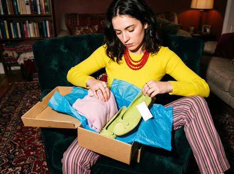 Focused young woman sitting in a dark green velvet chair while carefully unwrapping new clothing and green suede ballet flat shoes from a cardboard delivery box