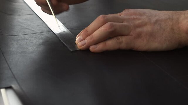 Leather craftsman marking a line on black leather using a metal ruler and an awl for a bespoke handcrafted project in a workshop