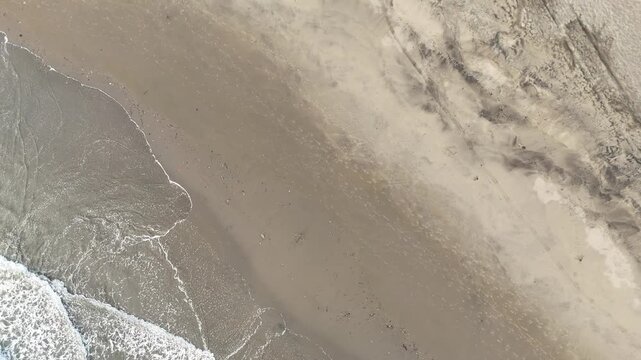 Zoom out aerial top view of sea waves hitting the beach. Overhead drone reveal shot of the ocean water, white foam, and sandy coastline directly from above.