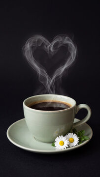 Romantic scene of a hot cup of coffee in a pale green ceramic cup with white daisy flowers on the saucer and steam rising to form a heart shape on a solid black background
