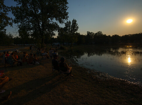Spain - 12 August 2026: View of people sitting on folding chairs by a lake at sunset with golden light reflecting on the water and trees in the background.