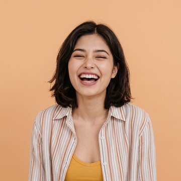 A joyful young Asian woman with a bob haircut, laughing with her eyes closed, wearing a striped shirt over a yellow top against a warm beige background.