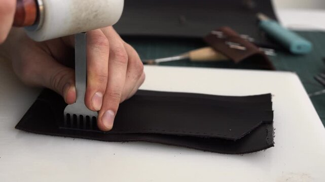 Craftsman punching stitching holes in black leather using a mallet and a pronged chisel in a workshop