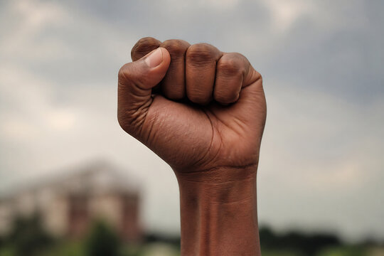 A close-up of a dark-skinned fist clenched in a determined gesture against a cloudy, blurred outdoor background.