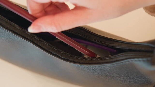 Close-up view showing hand guiding slim pencil into partially unzipped textured leather pouch, with zipper slider shifted aside, metal teeth exposed and purple lining visible inside