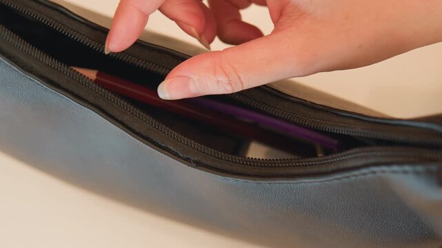 Close-up hand positions wooden-handled makeup brush into dark leather zipper pouch with purple lining, showing careful storage, partial opening, fingers on handle, warm light, and tactile material