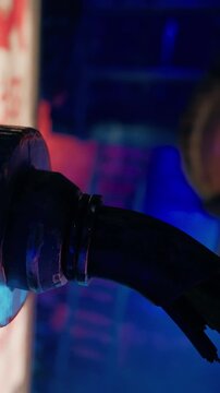 A fuel nozzle is shown close up at a gas station. Bright lights create colorful reflections and shadows around the area. The scene captures the busy atmosphere of fueling vehicles at night.