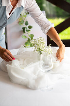 A woman in a blue vest and white shirt decorates a wedding table with white tulle fabric, flowers, and glasses.