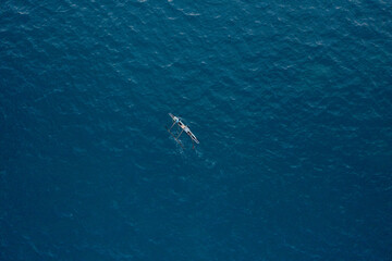 Minimalist top-down aerial shot of a traditional wooden outrigger fishing boat drifting alone in the vast, textured deep blue ocean water. © AndreyZayats