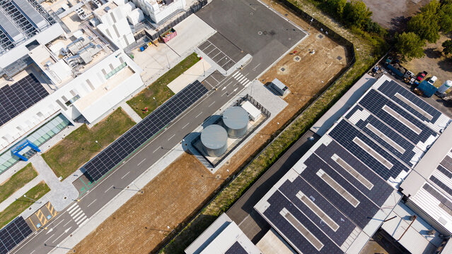 Aerial view of a modern data center facility featuring large industrial cooling tanks, solar panel arrays on rooftops, and paved access roads in Melegnano, Lombardy, Italy.