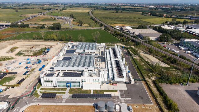 Aerial view of a modern data center facility with rooftop solar panels and cooling infrastructure, situated in an industrial area near Melegnano, Lombardy, Italy.