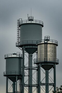 Tall metal water towers stand against cloudy sky. Industrial storage tanks provide fluid supply for local town infrastructure. Large grey elevated reservoirs feature ladders plus railings for