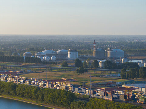 Aerial view of an industrial port facility with large spherical and cylindrical storage tanks, rows of shipping containers, and gantry cranes along a canal in the Netherlands.