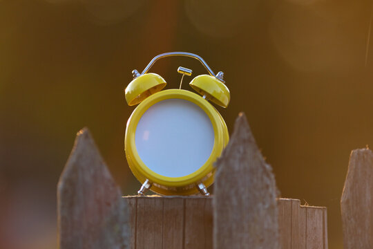 A bright yellow retro alarm clock is standing on an old wooden fence. Instead of a dial, there&rsquo;s an empty space for text.