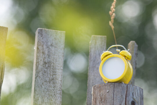 A bright yellow retro alarm clock is standing on an old wooden fence. Instead of a dial, there&rsquo;s an empty space for text.