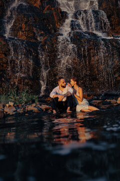 Romantic couple sitting near waterfall with water reflection, young man and woman looking at each other at sunset
