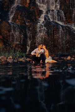 Passionate couple kissing sitting near waterfall with water reflection, romantic lovers on rocky shore at sunset