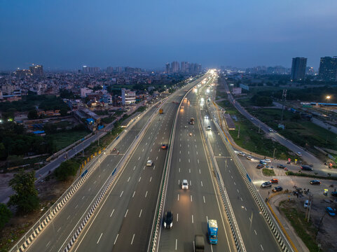 Aerial view of a multi-lane highway at dusk with cars driving under streetlights and a distant city skyline in Gurgaon, Haryana, India.