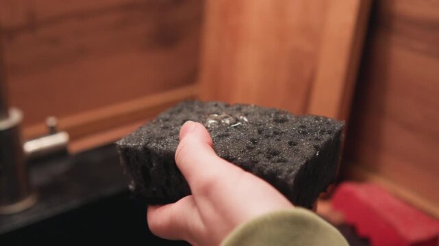 Right hand holds textured dark sponge above sink, squeezing out water while soap foam and droplets gather in perforations; visible dripping motion and glossy fingertips suggest routine cleaning