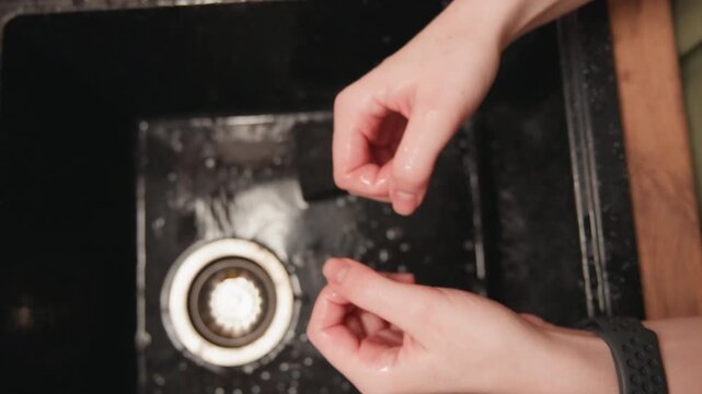 Top-down close-up shows hands removing debris from dishwasher filter beneath running water inside dark appliance cavity, with droplets, splashes, careful gripping, and routine kitchen maintenance