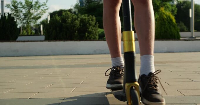 Close up of child feet riding scooter through sunny urban park. Moving sneakers and wheels capture active childhood, outdoor recreation, mobility and summer lifestyle during leisure time.