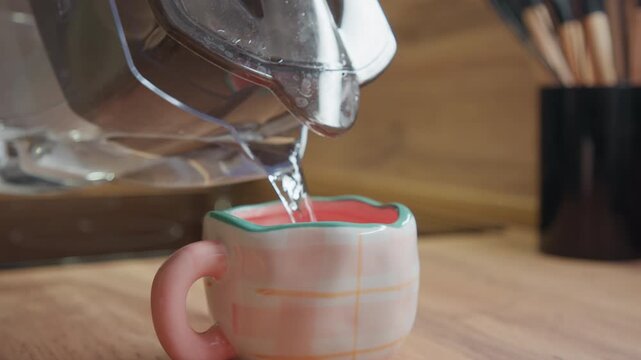Close-up of clear glass pitcher tilted to pour steady stream of water into pastel ceramic mug on wooden countertop; visible thin splash and ripple at mug rim, green interior glaze with pink liquid