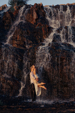 Passionate couple embracing under waterfall, strong man holding woman in arms standing near dark wet rocks at sunset