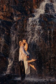 Passionate couple embracing under waterfall, strong man holding woman in arms standing near dark wet rocks at sunset