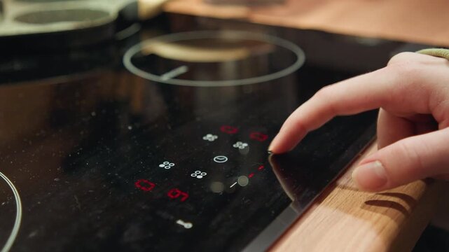 Adult hand presses illuminated touch control on glossy black induction cooktop, with glowing red and white icons, reflective ceramic surface, and modern kitchen setting suggesting cooking adjustment