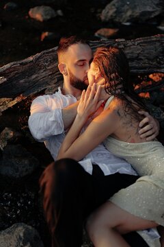 Passionate couple kissing lying together on beach leaning on dry log at sunset