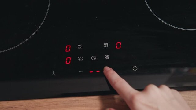 Close-up low-angle view of fingertip pressing illuminated touch controls on glossy black induction cooktop, showing glowing red numeric display and icons while adjusting heat, timer, and cooking