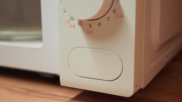 Close view of hand turning microwave timer dial while fingertips press lower control panel on white plastic casing set above wooden countertop, showing simple kitchen adjustment under warm indoor