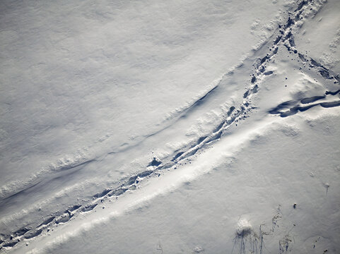 Aerial top-down view of solitary human footprints cutting a distinct path across a vast, pristine white snow-covered field during a crisp winter day, creating a journey concept