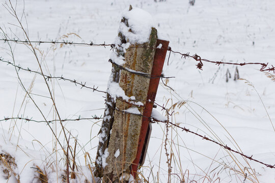 Weathered concrete post wrapped in rusty barbed wire stands solitary in a desolate snow covered field, capturing the stark beauty of a quiet, freezing winter day in the countryside