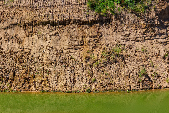 Detailed view of a textured clay and sand quarry wall with layered geological formations rising above a calm green pond surface in a bright sunny summer day