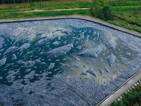 Aerial view of a sewage sludge storage facility featuring a large basin covered with a dark protective membrane for odor control, surrounded by green vegetation in Riga, Latvia.