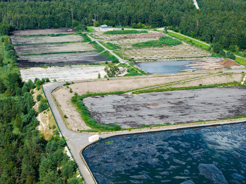 Aerial view of a sewage sludge storage facility featuring a large black basin covered with a membrane for odor control, surrounded by forest and access roads in Riga, Latvia.