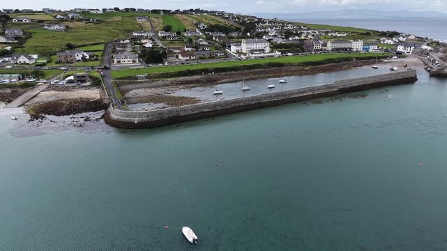 Drone flying across the beautiful Irish blue bay, County Sligo, Ireland, showcasing a stone breakwater and a pier, a numerous sailing boats moored along the pier and a shoreline. The smooth cinematic