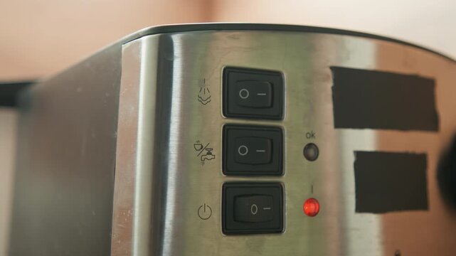 Horizontal close-up shows stainless steel toaster control panel with three black rectangular buttons, fingertip pressing one button and small red indicator light glowing on metal surface