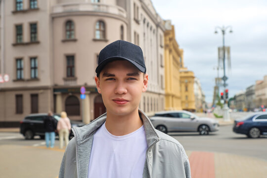 Young man in light clothes and black cap standing on wide city street, enjoying urban lifestyle and casual fashion during summer day outdoors.