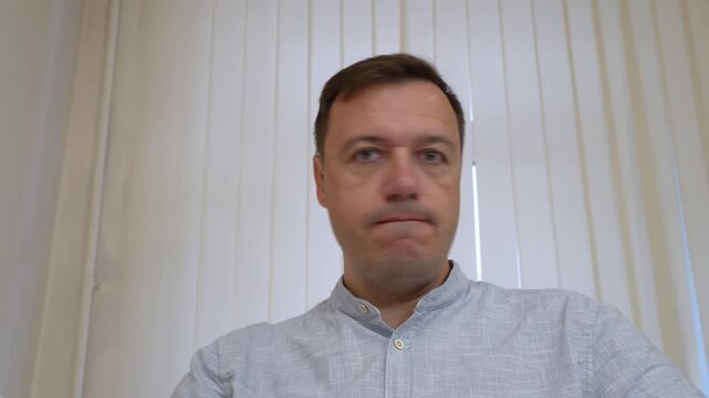 Caucasian adult man in casual shirt maintains direct gaze at camera, subtly opening his mouth in gentle expression of surprise or reaction while indoors.