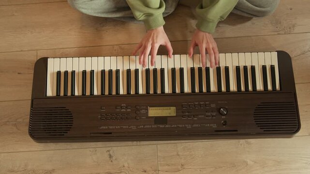 Top-down scene showing person seated on wooden floor playing portable electronic keyboard, both hands pressing cream keys with gentle finger movement, casual home setting, warm light, focused posture