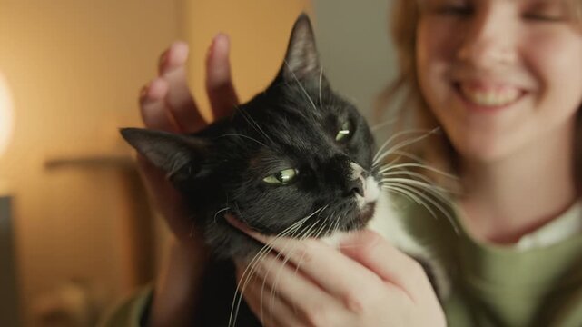 Black and white tuxedo cat rests against person's chest while caring hands support chin and neck, creating subtle head turn, half-closed green eyes, relaxed fur, and warm domestic intimacy