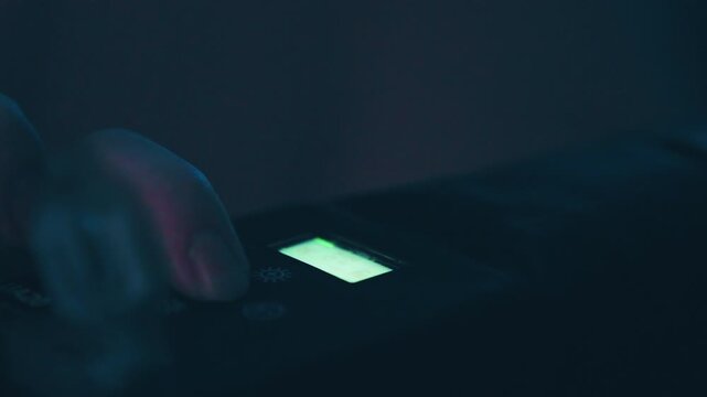 Fingertip applies deliberate downward press to illuminated rectangular button on dark handheld electronic device, with soft green backlight, cool blue shadows, shallow focus, and moody low-key