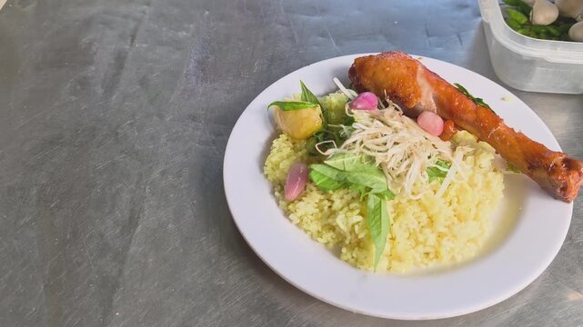 Hand placing a plate of authentic Phu Yen style chicken rice with roasted chicken leg, pickled whole shallots and butter on stainless steel table